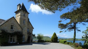Un bâtiment en pierre doté d’une tour, entouré d’une allée de gravier et d’une végétation luxuriante.