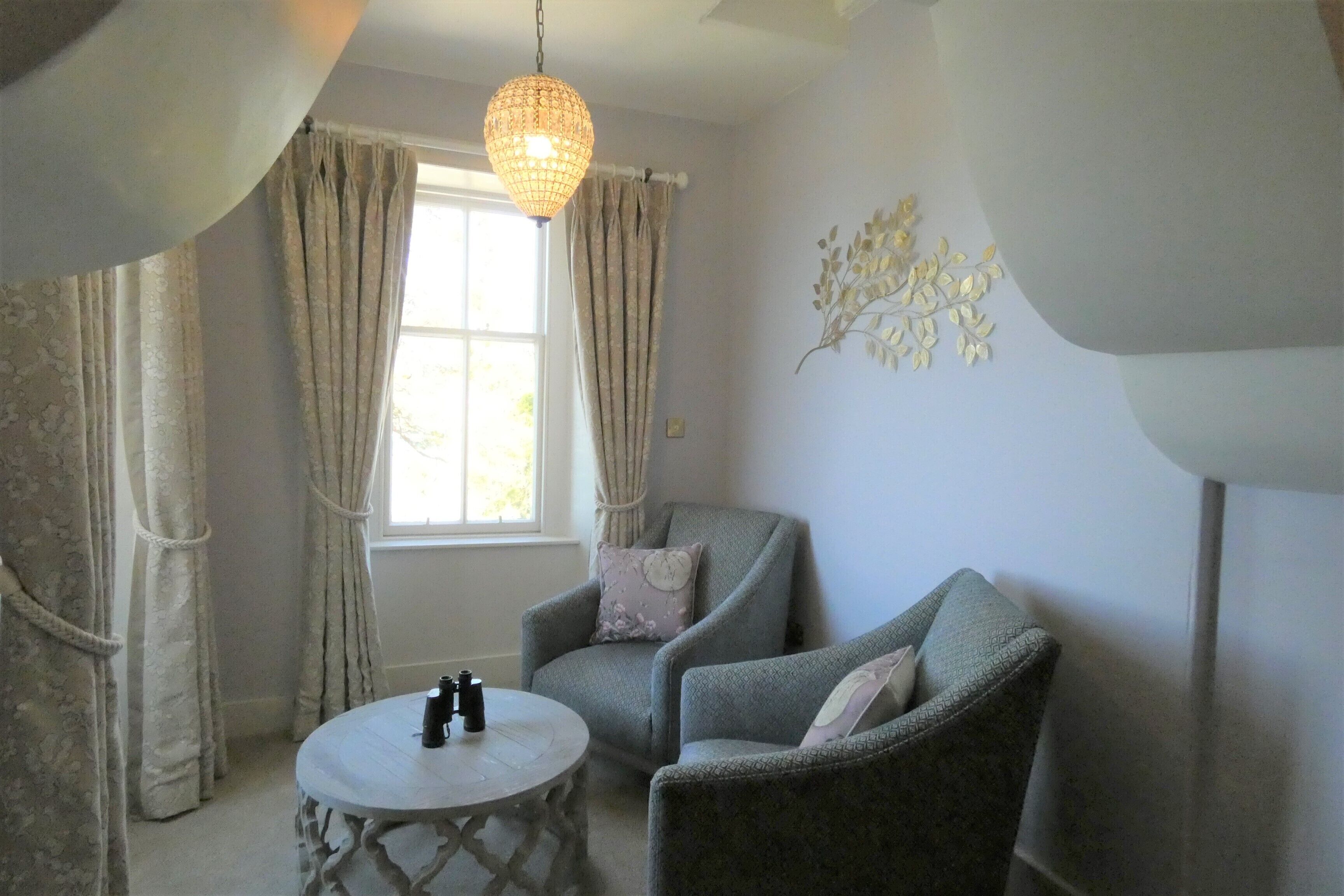 A room with two grey armchairs, a round table with binoculars, a decorative wall piece, and a window with curtains.