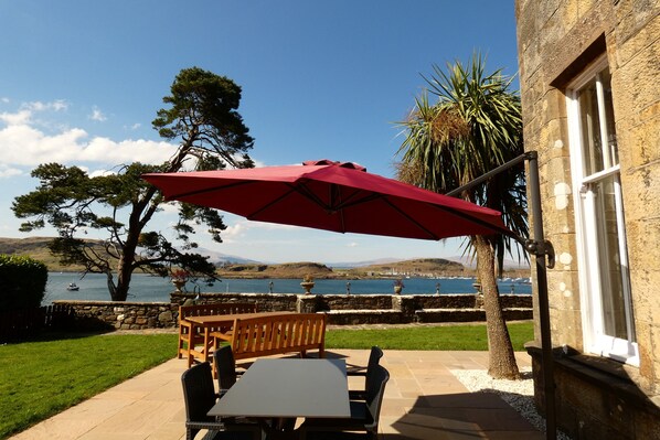 An outdoor patio with a table and chairs under a red umbrella, overlooking a body of water and hills. - Dungallan Country House (Oban)