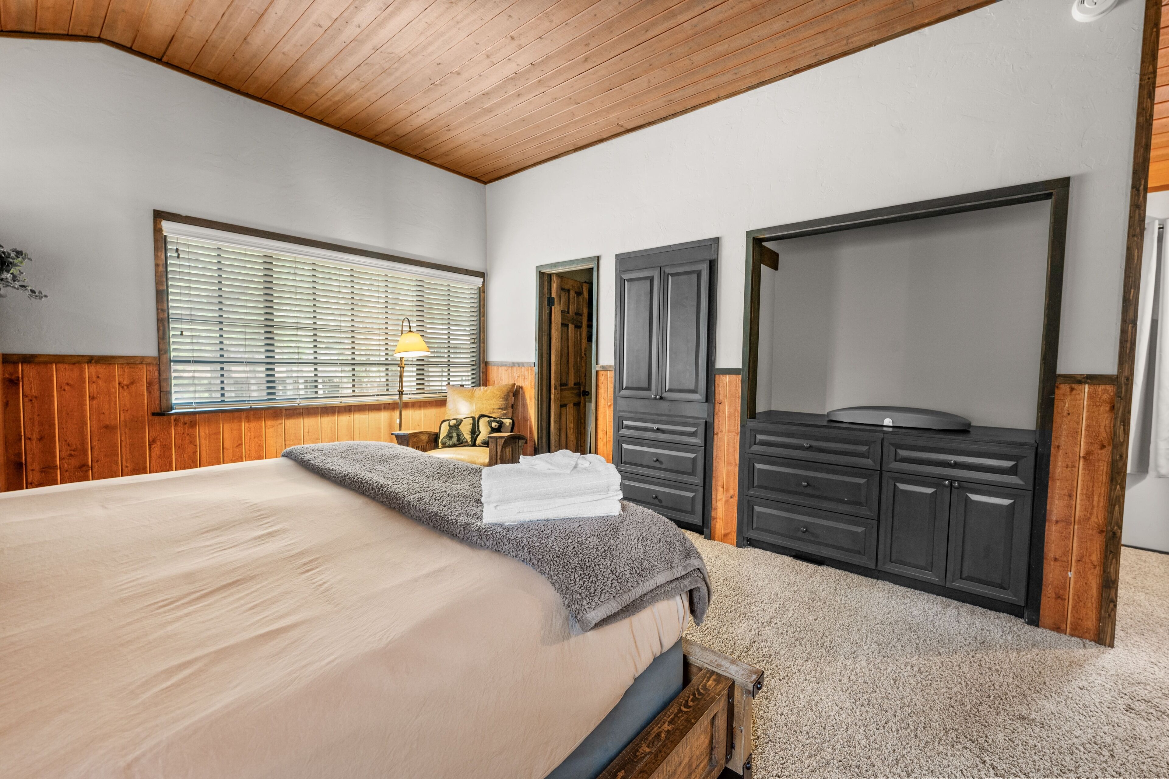 A bedroom with a large bed, wooden ceiling, a window with blinds, a bedside table with a lamp, and a wardrobe.