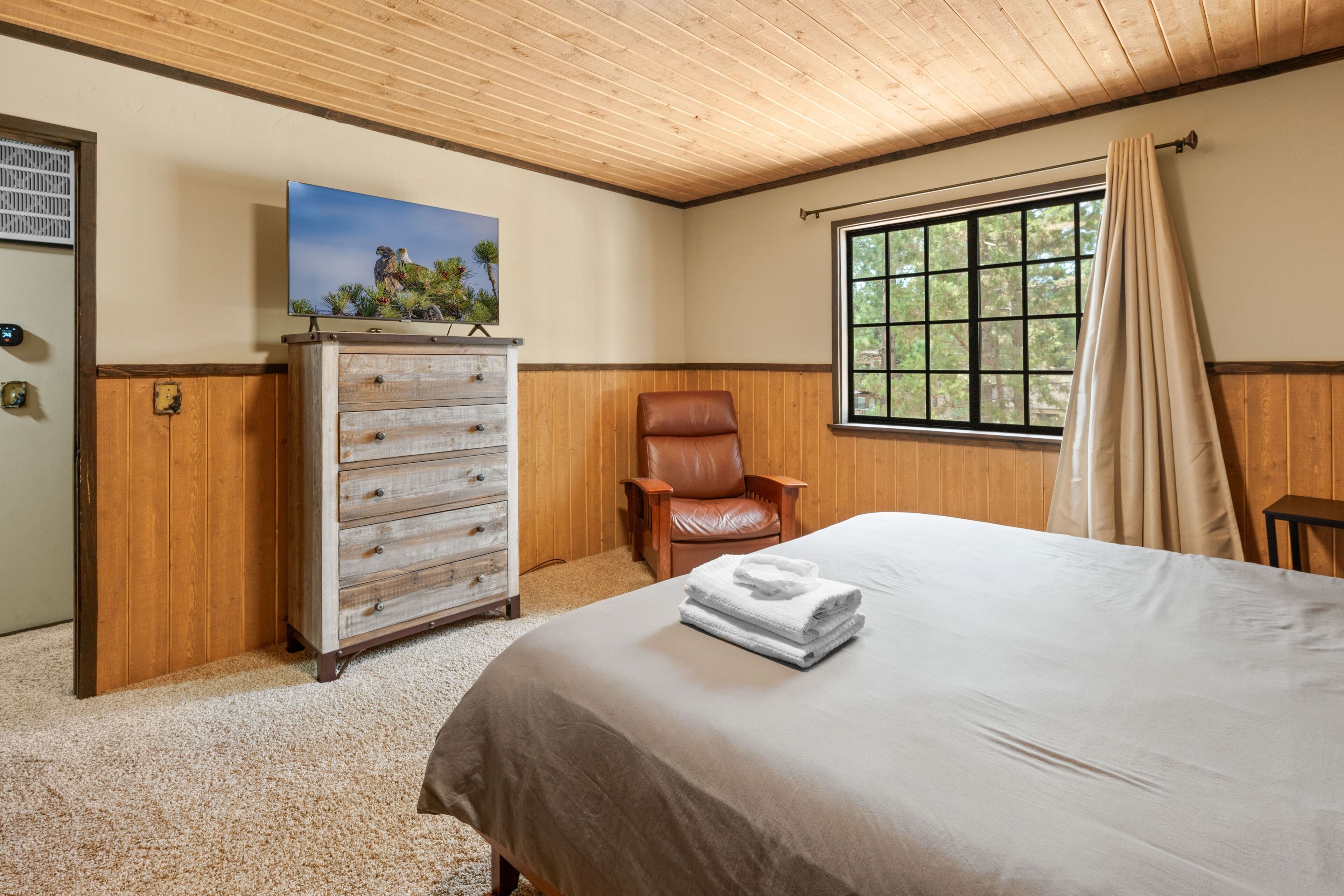 Un dormitorio con una cama, un aparador de madera, un sillón de cuero, una ventana con vistas a árboles y un cuadro en la pared.