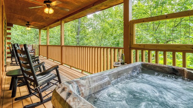 A wooden deck with a hot tub, rocking chairs, and a view of lush greenery. - Kozy Cub Cabin, Gated Resort, Pool Table (Sevier County)