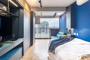 A modern bedroom with a large bed, a desk with a chair, a small stool, and a TV.