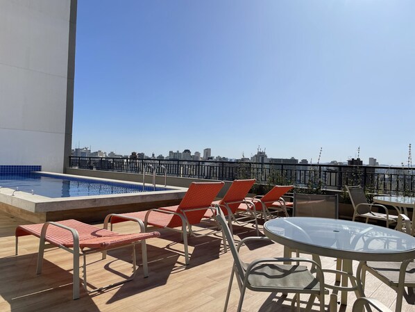 A rooftop pool area with lounge chairs and a glass table, overlooking a cityscape.