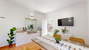 A modern kitchen with a dining area, a TV mounted on the wall, and a view of the outdoor patio. - Quinta Casalinho - T2 Amendoeira (Guia)