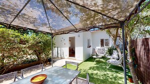 An outdoor patio with a table and chairs, a small garden area with artificial grass, and a white building with a door and windows. - Quinta Casalinho - T2 Amendoeira (Guia)