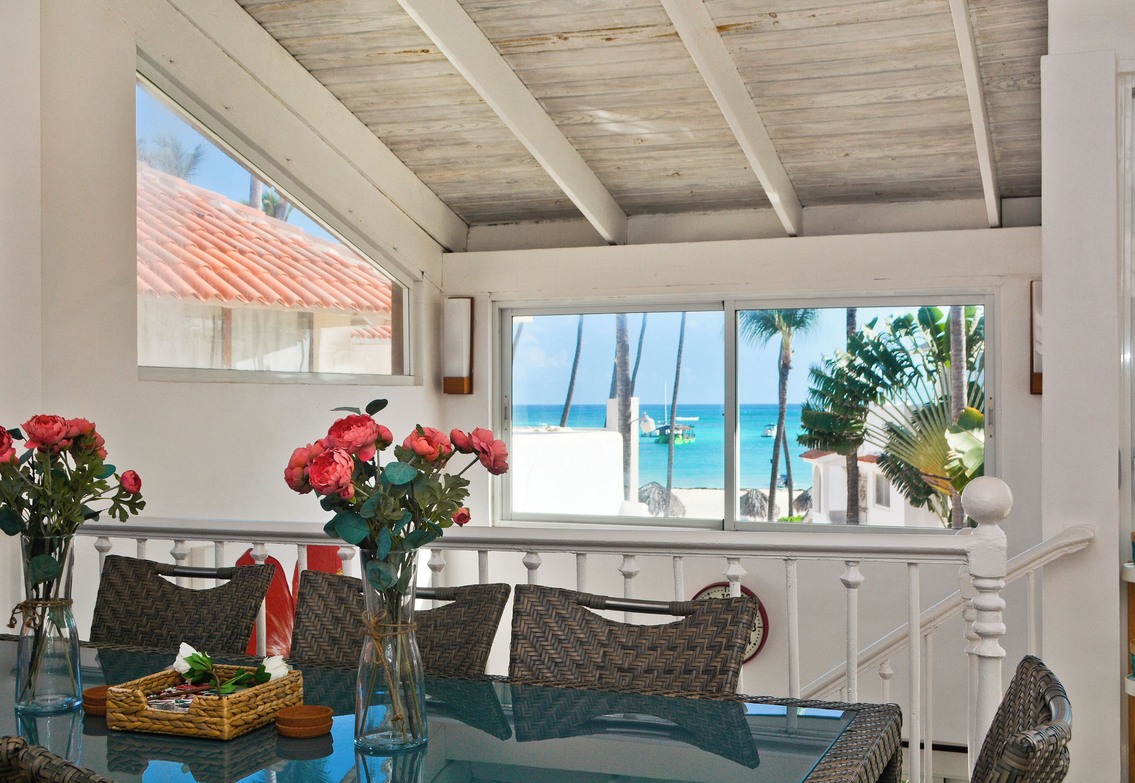 A dining area with a glass table, wicker chairs, and a view of the beach and ocean through a window.