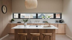 A modern kitchen with a large island, wooden stools, a hanging light fixture, and a view through the window.