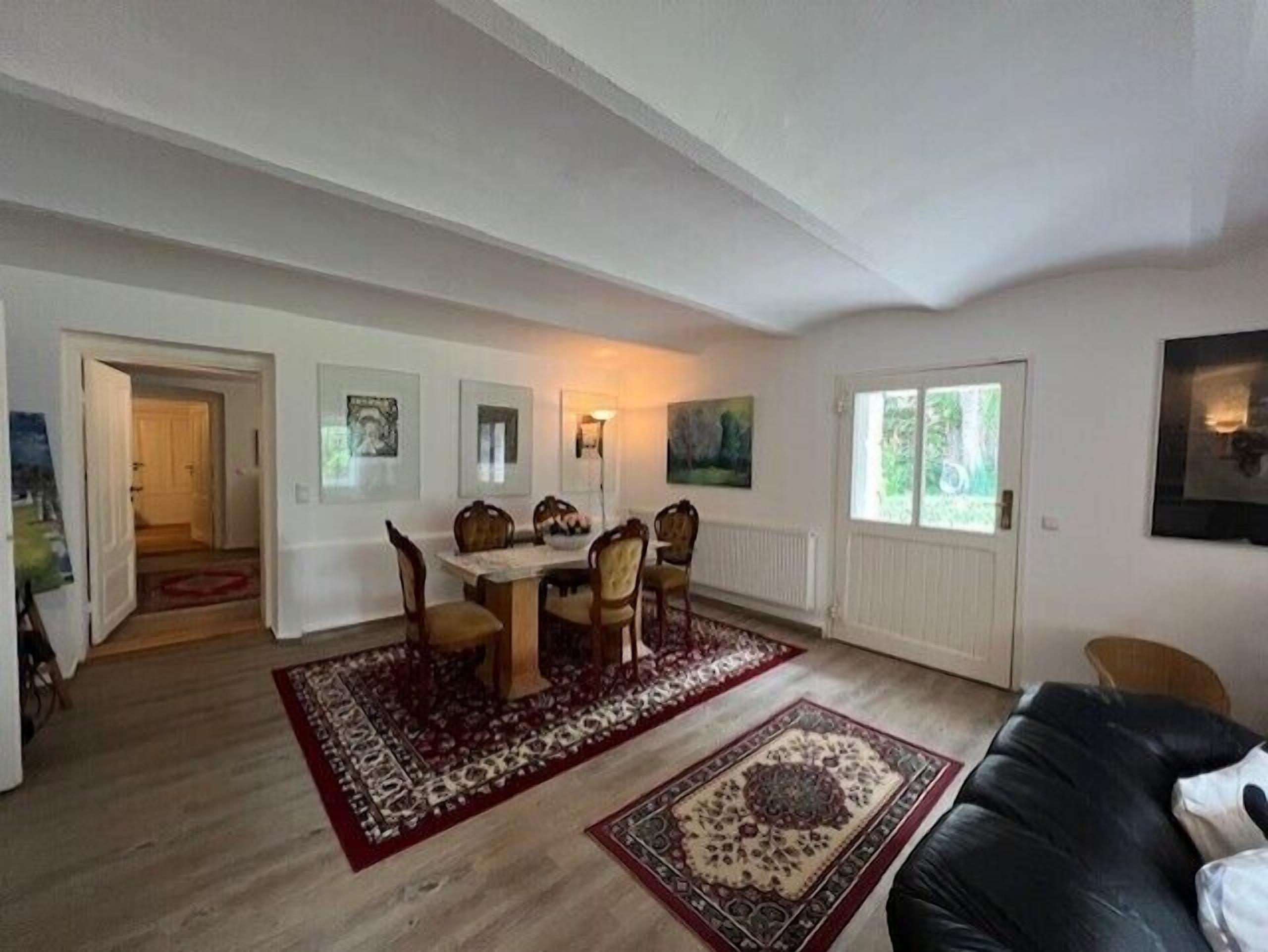 Un salon spacieux comprenant un coin repas, avec une grande table et des chaises en bois, un canapé en cuir noir et un tapis aux motifs rouges et beiges.