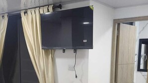 Habitació amb televisió de pantalla plana penjada a la paret, cortines beix, una porta de fusta i un paviment de rajoles.