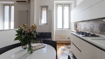 A modern kitchen with a gas stove, white cabinets, and a hexagonal tile backsplash. There is a dining table with chairs and a vase of flowers.