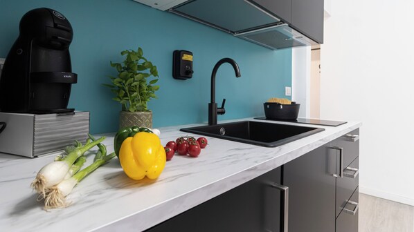 Une cuisine moderne avec un évier noir, une machine à café, une plante en pot et des légumes frais sur le plan de travail.