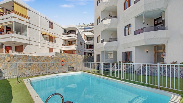 A swimming pool with a stone wall and metal railing, surrounded by multi-story buildings with balconies. - Apartment Two Minutes from the Beach. Puerto Rico (Mogán)