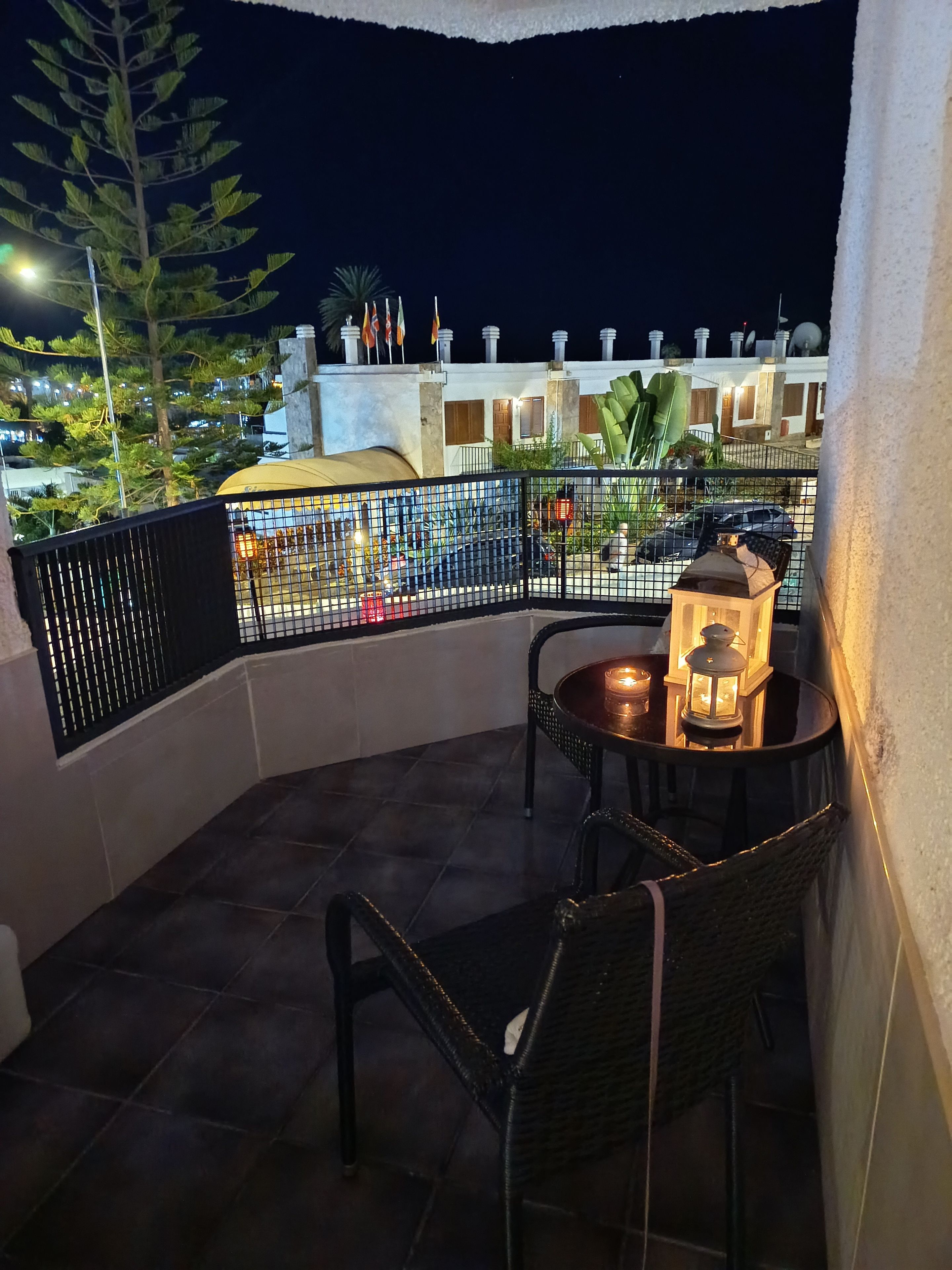 A balcony with a table and chairs, a lantern, and a view of buildings at night.