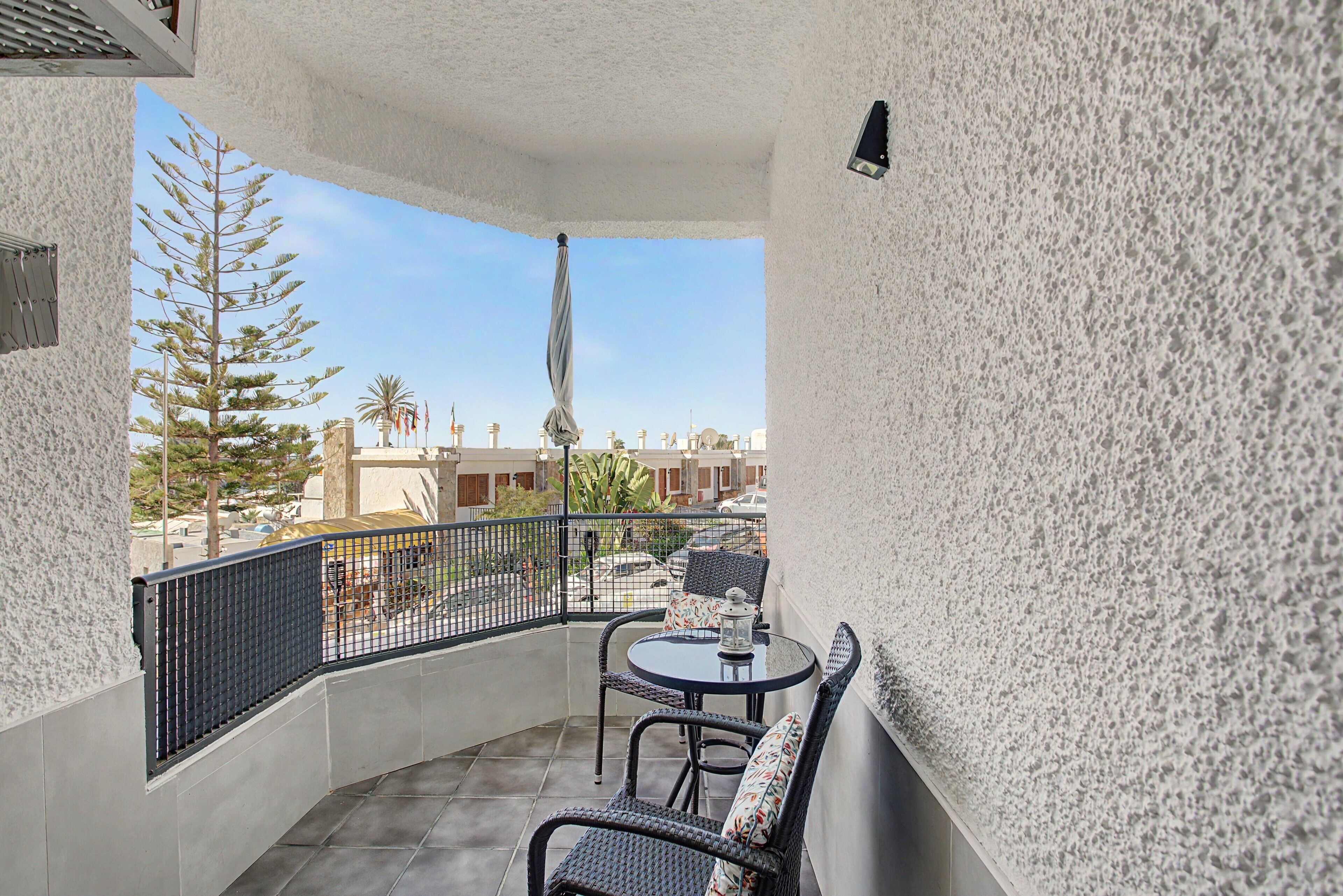 Un balcón con una mesa y sillas, un lámpara colgante y vistas de los edificios y palmeras circundantes.