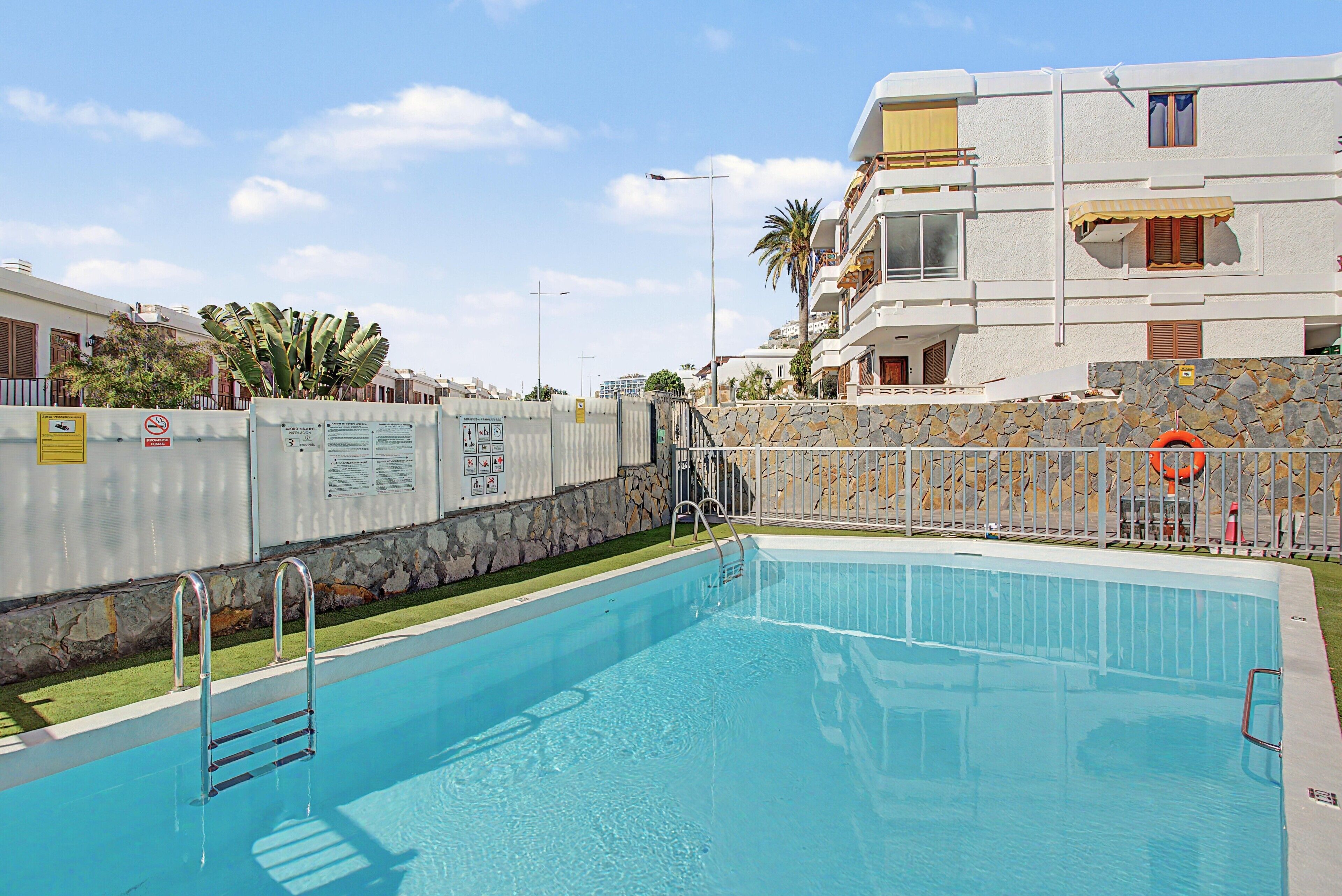 Una piscina con un muro de piedra y una valla de metal, rodeada de edificios y palmeras.