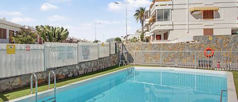 Una piscina con un muro de piedra y una valla de metal, rodeada de edificios y palmeras.