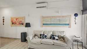 Un salon avec un canapé beige, un tableau représentant une plage et un climatiseur fixé au mur.