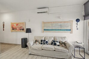 Un salon avec un canapé beige, un tableau représentant une plage et un climatiseur fixé au mur.