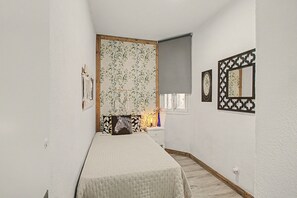Une chambre à coucher comprenant un lit, une commode, un miroir et un mur recouvert d’un papier peint à motifs floraux.