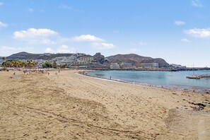 A sandy beach with a clear sky, a rocky hill in the background, and a body of water. - Apartment Two Minutes from the Beach. Puerto Rico (Mogán)
