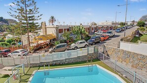 A swimming pool with a stone wall and metal railing, surrounded by parked cars and a row of buildings. - Apartment Two Minutes from the Beach. Puerto Rico (Mogán)