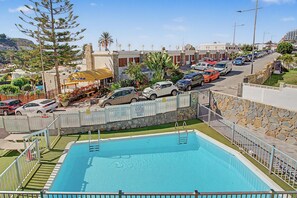 A swimming pool with a stone wall and metal railing, surrounded by parked cars and a row of buildings. - Apartment Two Minutes from the Beach. Puerto Rico (Mogán)