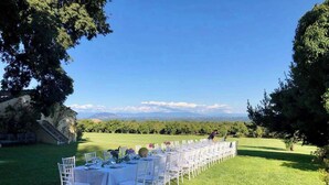 An outdoor dining area with white tables and chairs, set against a backdrop of a vineyard and distant mountains. - New Estate of Joy a Magnificent Historic Villa Near Rome on Glorious Ground Pool and Olives Galore (Ronciglione)