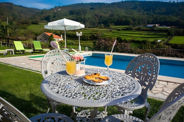 A poolside table with a plate of pastries, two drinks, and a white umbrella.