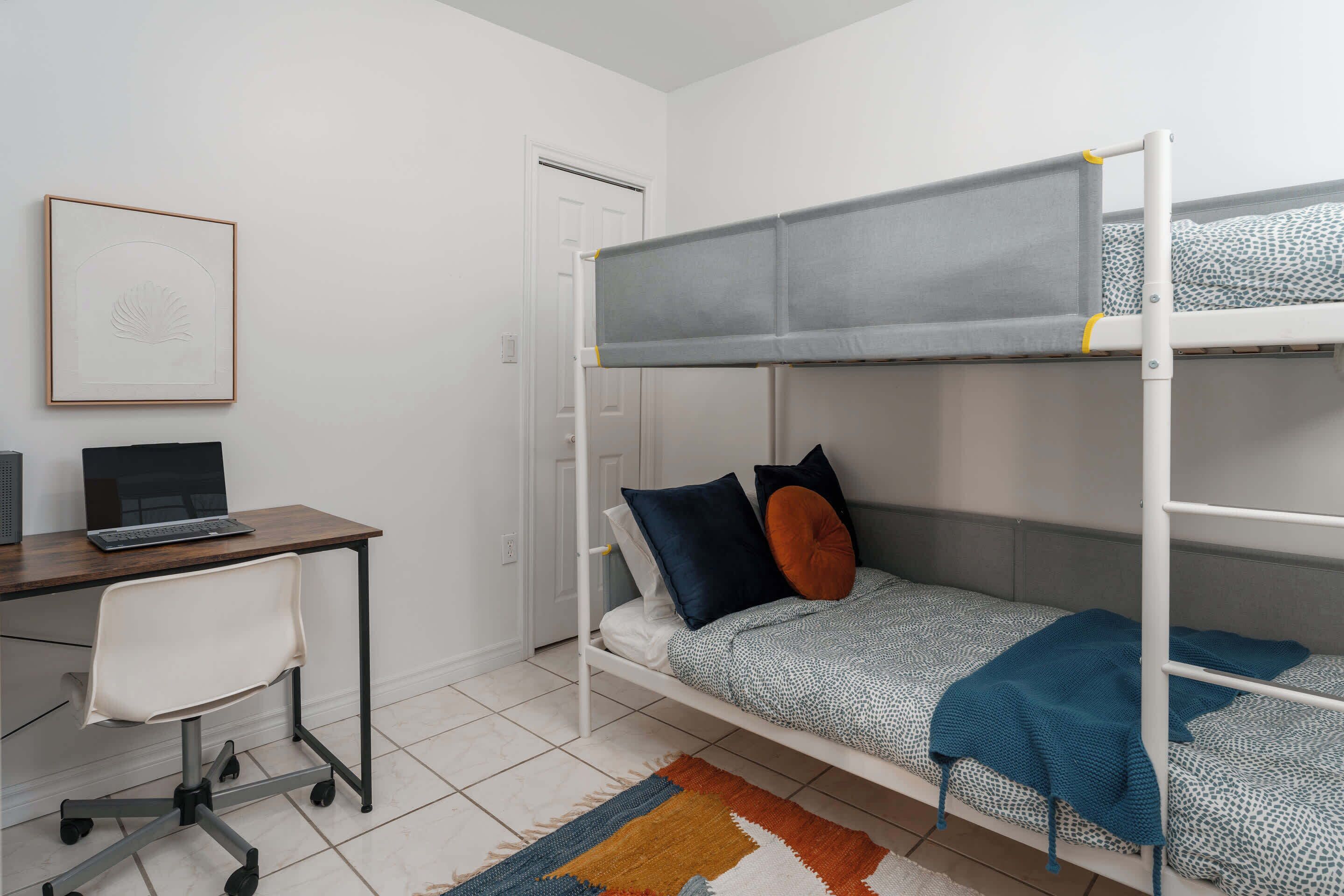 A dormitory room with bunk beds, a desk with a laptop, and a chair.