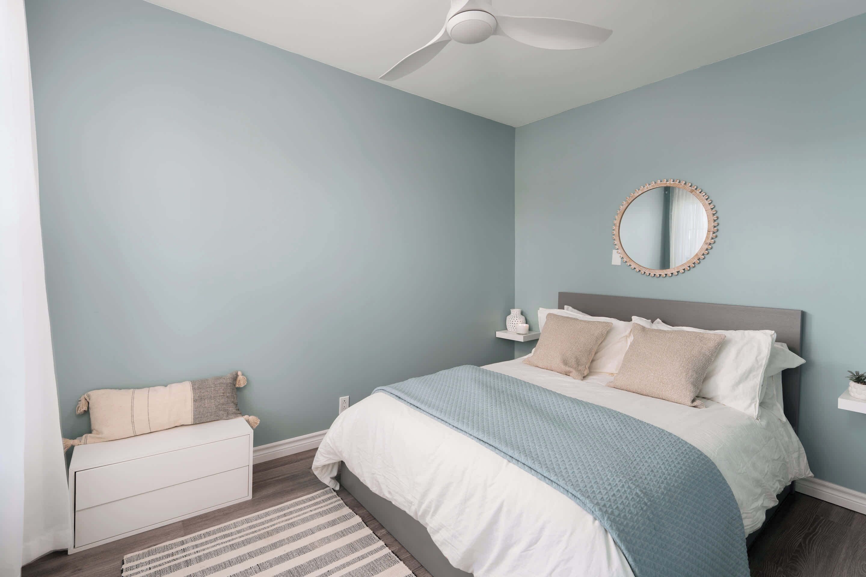 Quarto com cama, mesas de cabeceira, ventilador de teto, espelho redondo e parede azul clara.