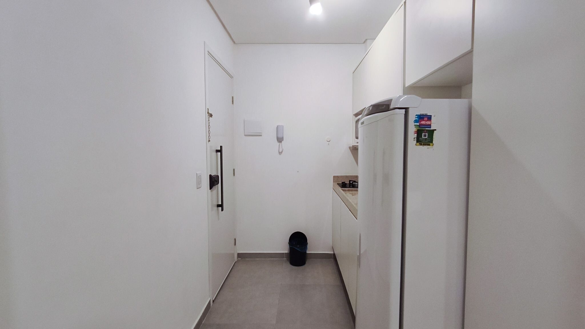 Un pasillo estrecho con un refrigerador, un bote de basura y una puerta con un teléfono en la pared.