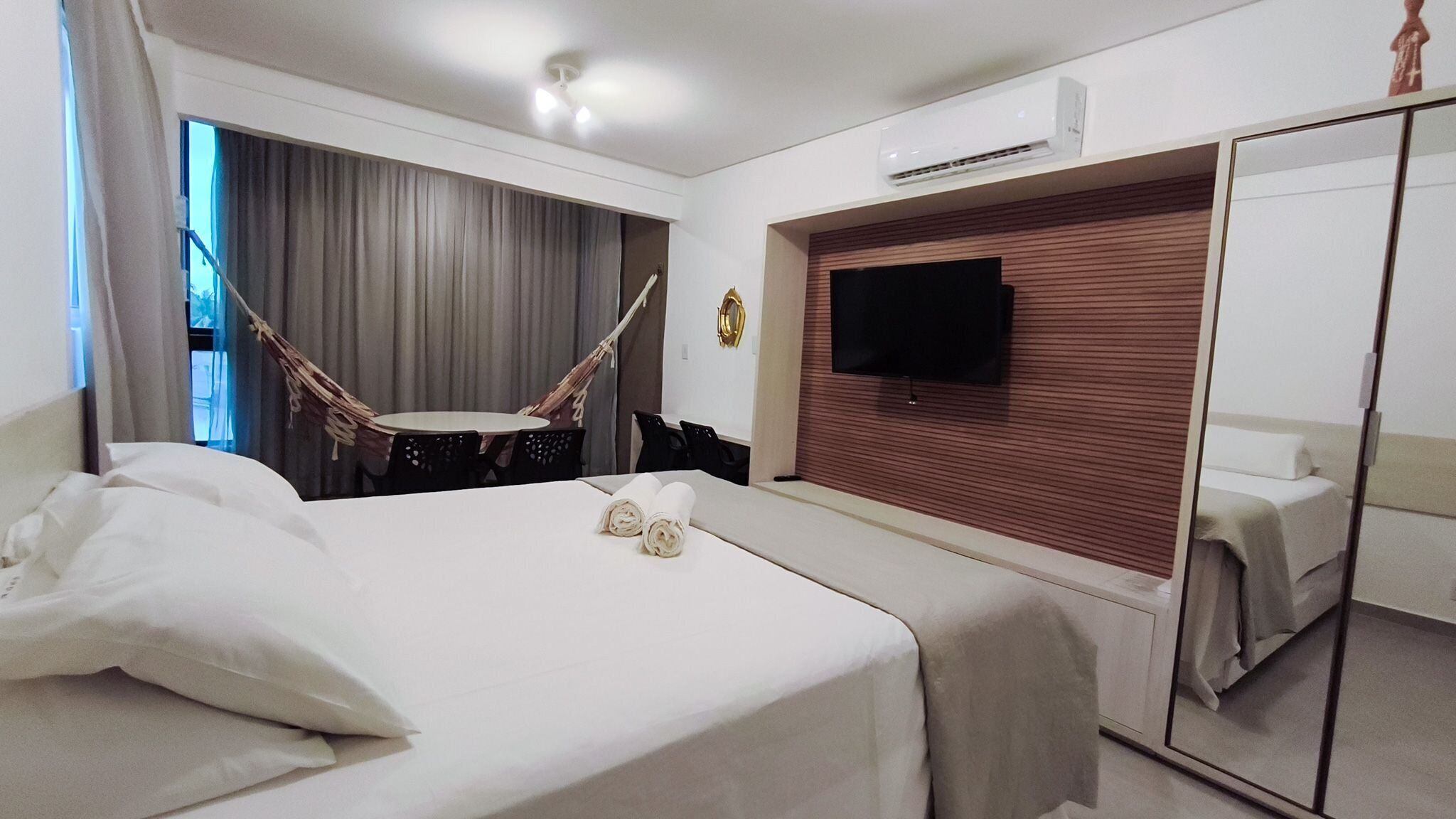 A hotel room with a bed, a TV mounted on the wall, a mirror, and a window with curtains.