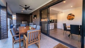 A modern kitchen with a large island, a dining area with a wooden table and chairs, and a sliding glass door leading to a patio. - Grange Retreat - Beach, BBQ (Grange)