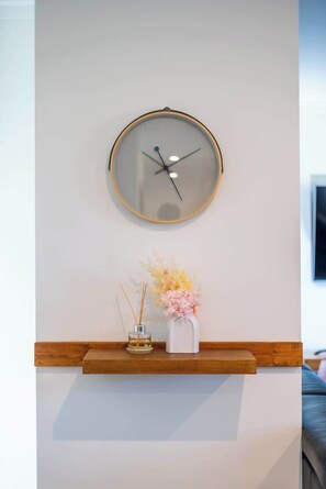 Eine wandmontierte runde Uhr mit Holzrahmen, darunter ein kleines Regal mit einer Vase mit getrockneten Blumen und einem Reed-Diffusor, und ein Fernseher zur Rechten.