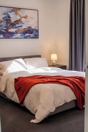 Una cama bien hecha con una colcha blanca y roja, una lámpara de noche y un cuadro en la pared.
