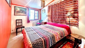 A bedroom with a large bed, a chair, a window with curtains, a hanging light fixture, and a tapestry on the wall.