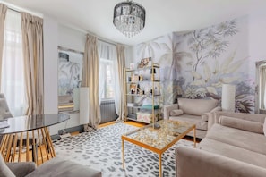 A living room with a glass coffee table, beige sofas, a glass dining table, a chandelier, a bookshelf, and a mural on the wall.