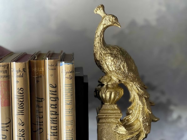 Una colección de libros con sus títulos visibles en el lomo, y una escultura de un pavo real dorado sobre un pedestal.