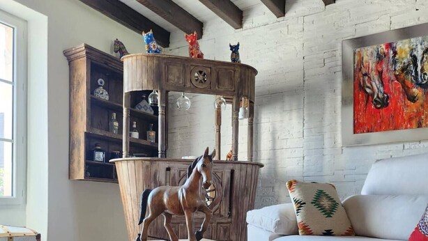 A rustic living room with a wooden coffee table featuring a horse sculpture, a white sofa with patterned cushions, and a wooden cabinet with glassware. - Domaine la Heroica (Maisonnais)
