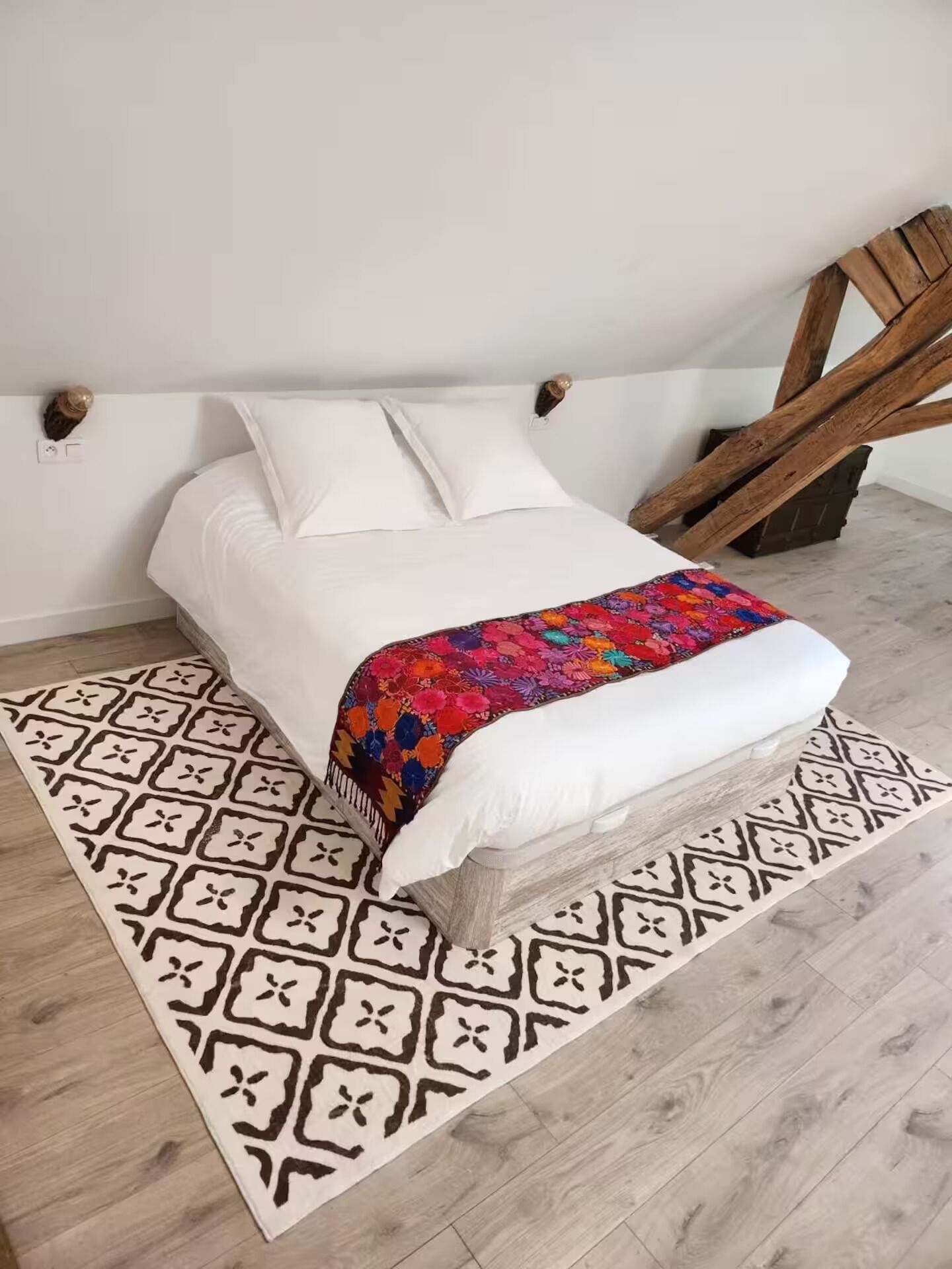A bed with a white mattress and pillows, a colorful blanket, and a patterned rug on a wooden floor.