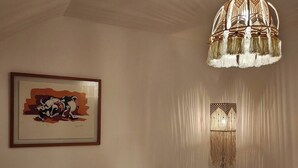 Une chambre à coucher comprenant un lit, une œuvre d’art encadrée, une lampe suspendue, une lampe sur pied et un petit tabouret en bois.