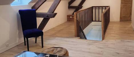 A loft-style room with a sloped ceiling, wooden beams, a glass coffee table, a blue chair, a leopard print rug, and a staircase.