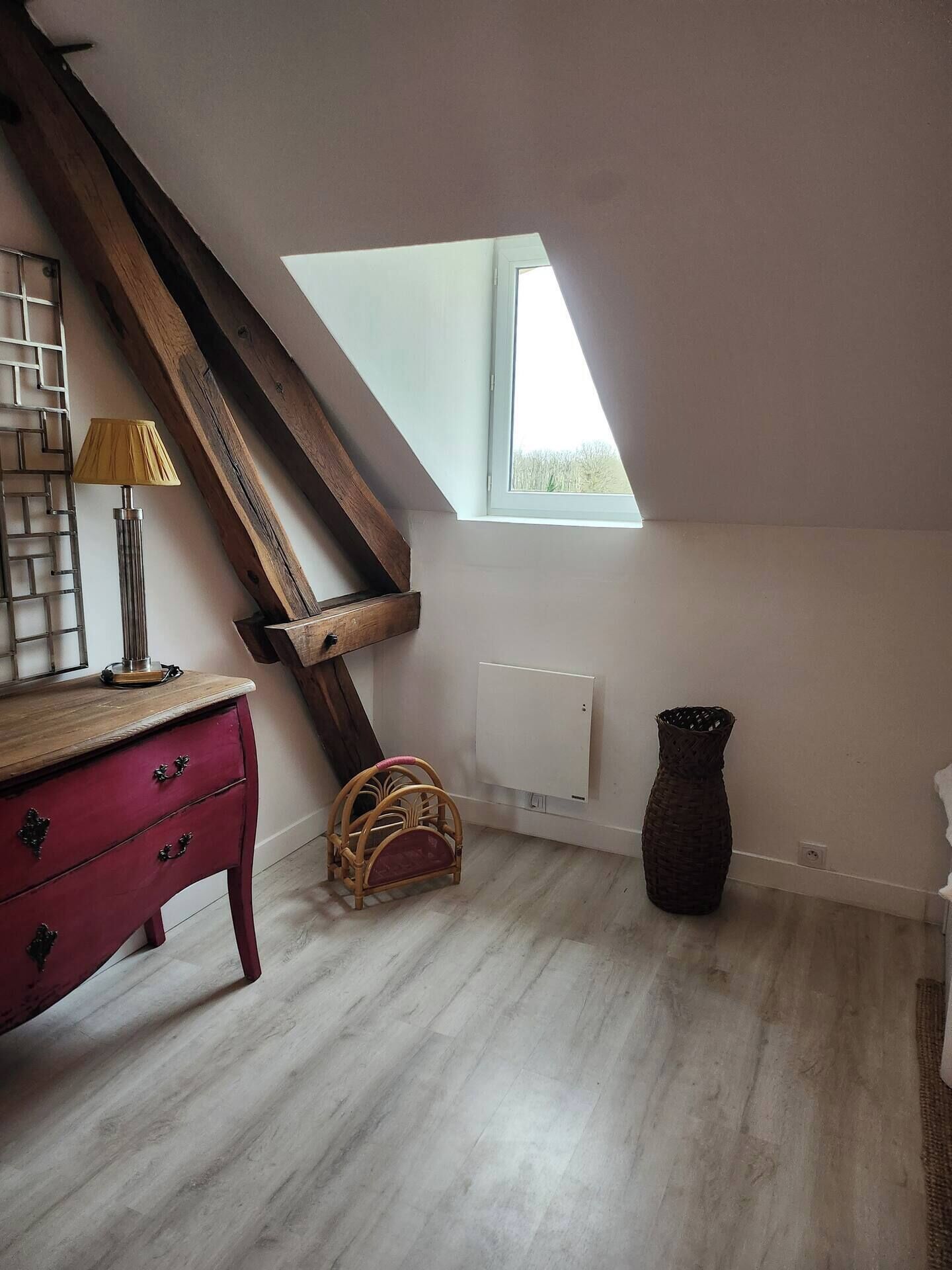 A room with a sloped ceiling, a wooden beam, a window, a radiator, a wicker basket, and a red dresser.