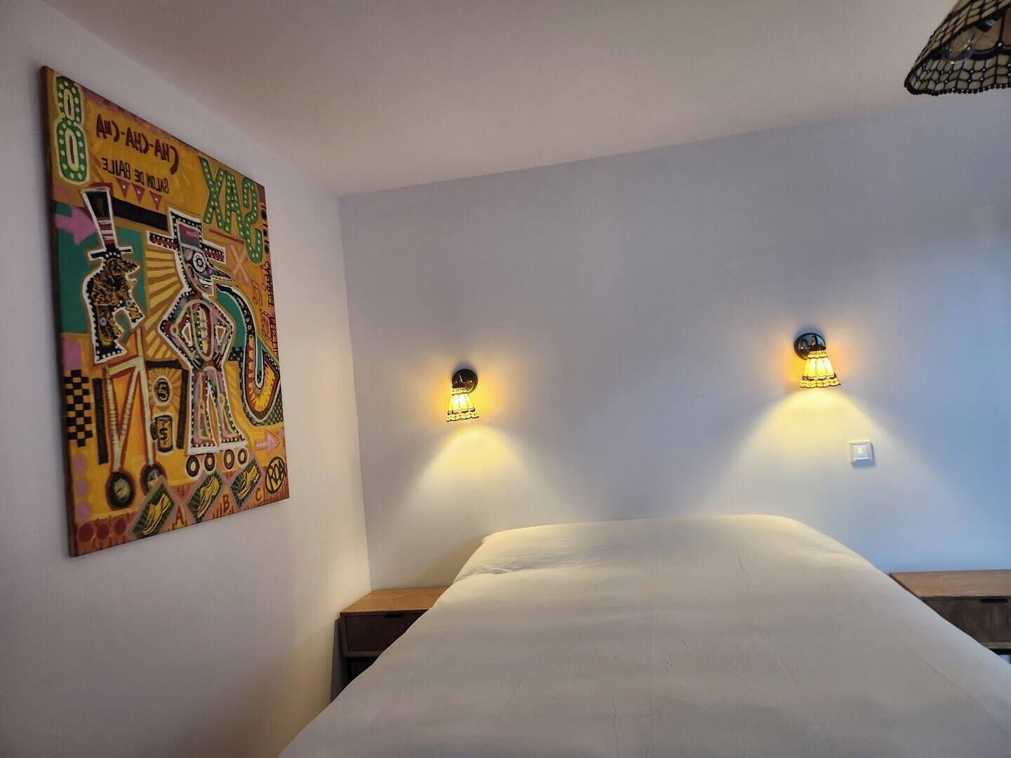 Une chambre d’hôtel avec un lit, deux lampes fixées au mur et un tableau abstrait aux couleurs vives accroché au mur.
