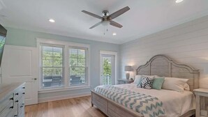 Une chambre à coucher comprenant un lit, des tables de chevet, un ventilateur de plafond et de grandes fenêtres équipées de stores.