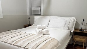 A neatly made bed with rolled towels, a bedside table with a lamp, and a window with blinds. - Charming apartment near Manhattan. Perfect for a getaway in NYC (North Bergen)