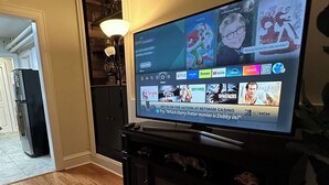A flat-screen TV mounted on a black entertainment center in a room with wooden flooring and a kitchen in the background. - Charming apartment near Manhattan. Perfect for a getaway in NYC (North Bergen)