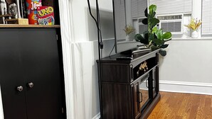 A room with a wooden floor, a black cabinet, a desk with a lamp, and a window with blinds. - Charming apartment near Manhattan. Perfect for a getaway in NYC (North Bergen)