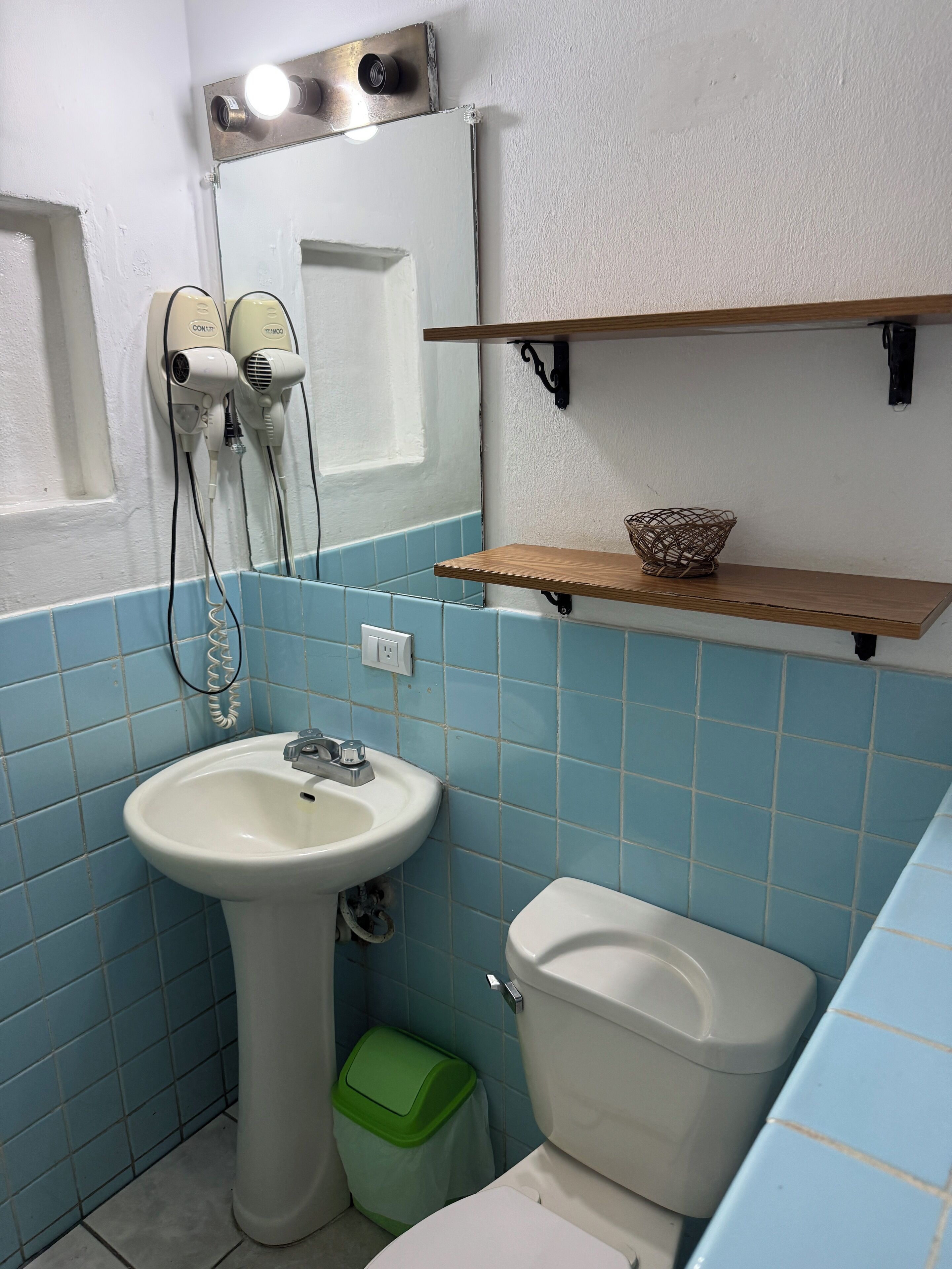 A bathroom with a sink, mirror, hair dryers, a toilet, and a green trash bin.
